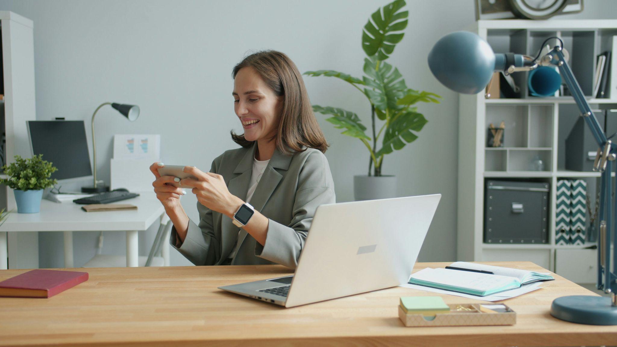 Mujer sonriente trabajando en oficina moderna mientras usa su teléfono móvil junto a una laptop y documentos sobre el escritorio.