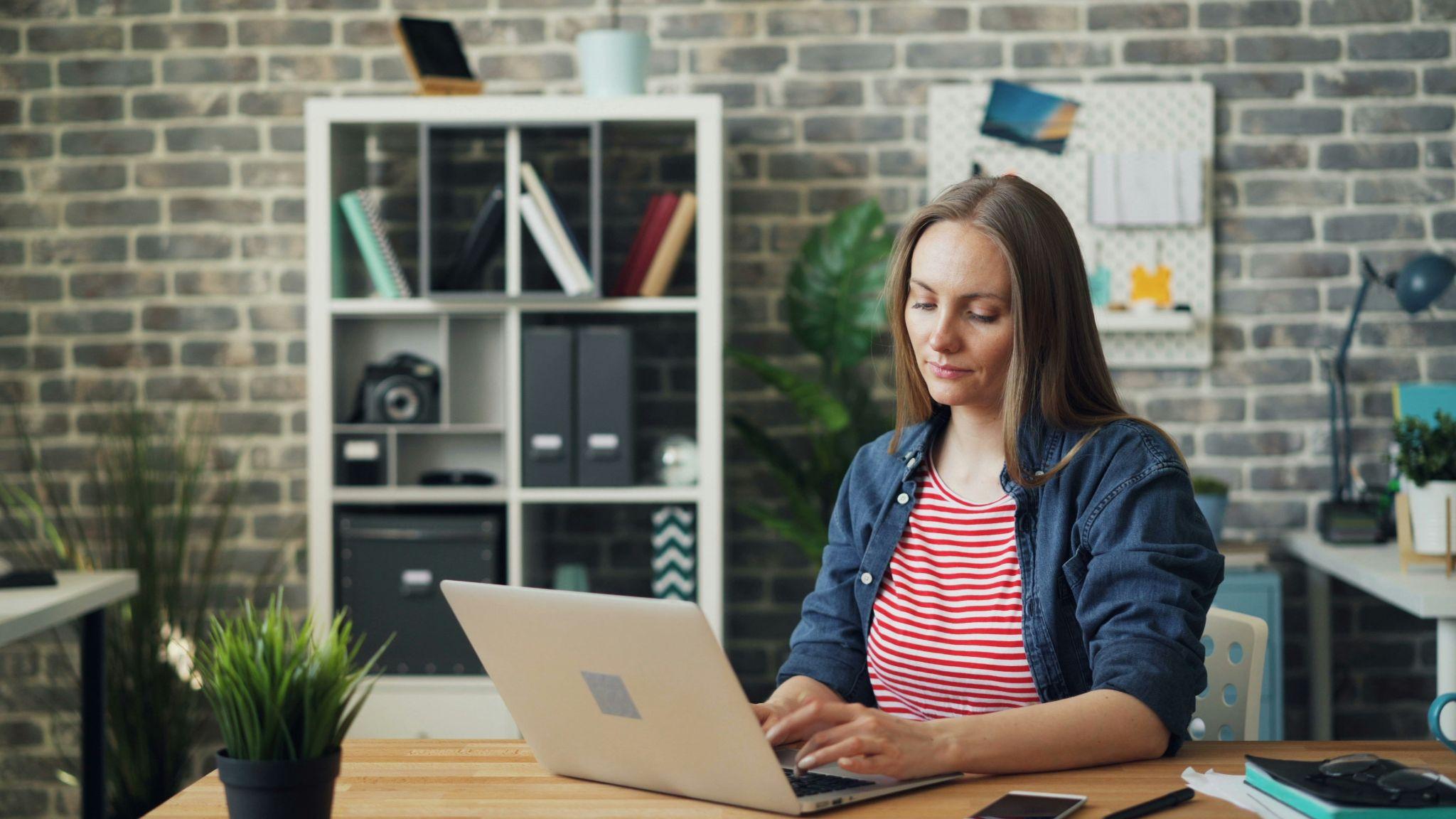 Mujer trabajando en su laptop en una oficina moderna con paredes de ladrillo y decoración minimalista.