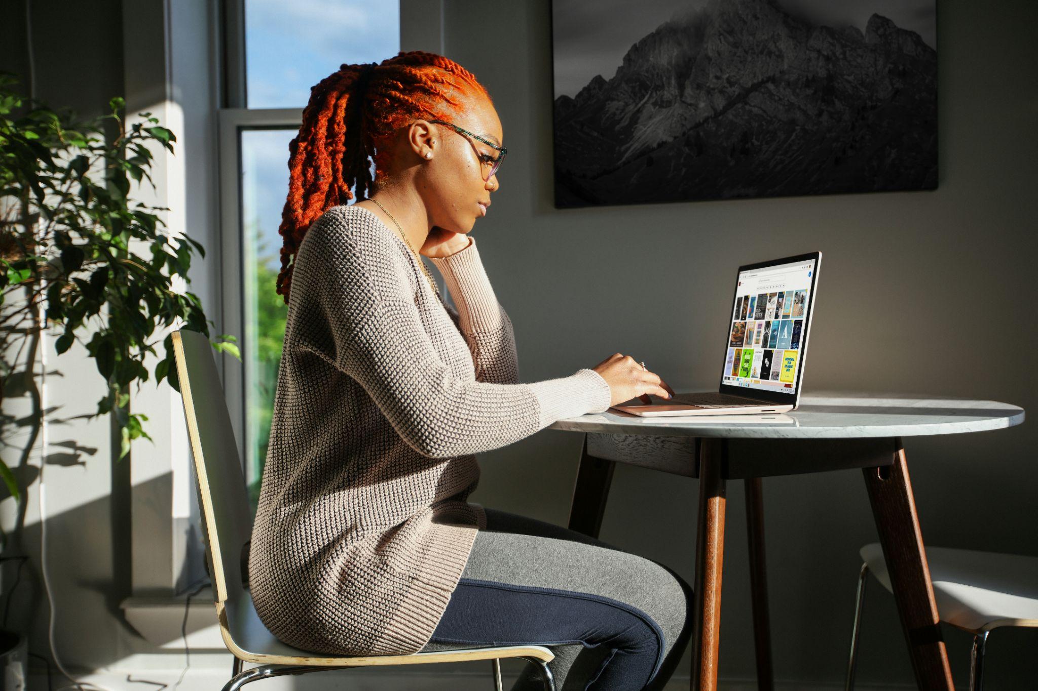 Mujer trabajando en su laptop junto a una ventana, concentrada en un entorno con luz natural y decoración minimalista.