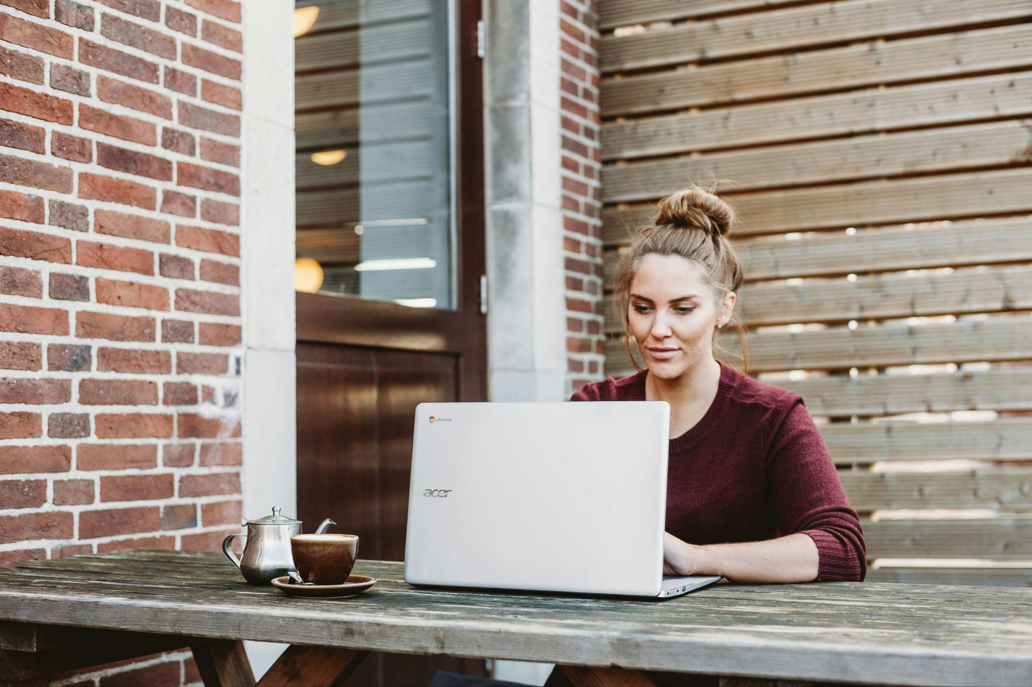 Mujer trabajando en laptop al aire libre en una terraza de café, con una taza y tetera sobre la mesa.